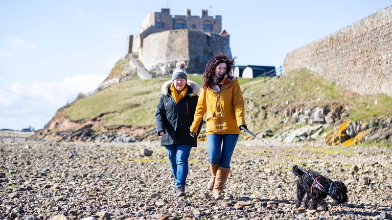 Two visitors walk along a pebbly beach with their black dog on a lead. In the background, a castle can be seen on top of the cliffs.